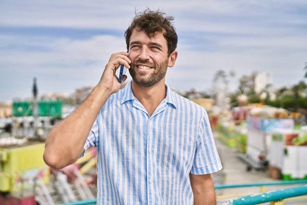Молодой латиноамериканец улыбается счастливо разговаривая по смартфону в парке аттракционов. - Фото, изображение