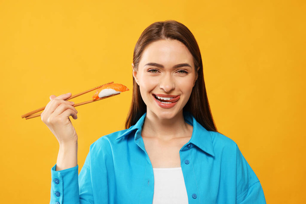 Happy beautiful young woman holding sushi with chopsticks on orange background - Photo, image