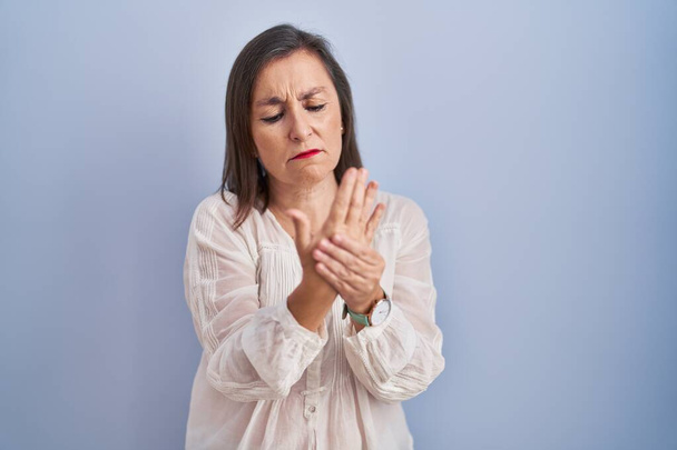 Латиноамериканка средних лет, стоящая на синем фоне, страдает от боли в руках и пальцах, воспаления артрита  - Фото, изображение