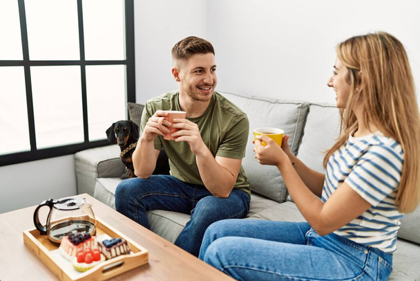 Junges hispanisches Paar lächelt glücklich beim Frühstück auf dem Sofa mit Hund zu Hause. - Foto, Bild