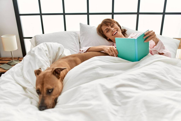 Młoda kaukaska kobieta czyta książkę leżącą na łóżku z psem w sypialni - Zdjęcie, obraz