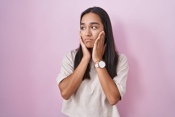 Junge hispanische Frau steht über rosa Hintergrund müde Hände, die Gesicht, Depression und Traurigkeit verdecken, verärgert und irritiert wegen des Problems  - Foto, Bild