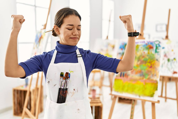 Young brunette woman at art studio showing arms muscles smiling proud. fitness concept.  - Photo, Image
