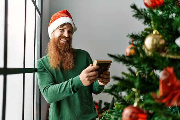 Young redhead man looking photo standing by christmas tree at home - Foto, imagen