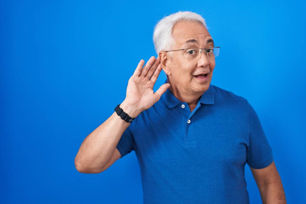 Een man van middelbare leeftijd met grijs haar op een blauwe achtergrond die glimlacht met hand op oor en luistert naar geruchten of roddels. doofheid.  - Foto, afbeelding