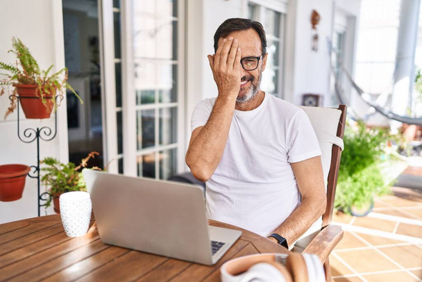 Muž středního věku používající počítač notebook doma zakrývající jedno oko rukou, sebevědomý úsměv na tváři a překvapivé emoce.  - Fotografie, Obrázek