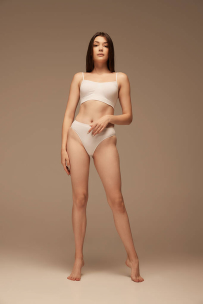 Portrait of young beautiful woman with sportive body posing in white underwear isolated over dark beige studio background. Concept of beauty, body and skin care, health, plastic surgery, cosmetics - Φωτογραφία, εικόνα