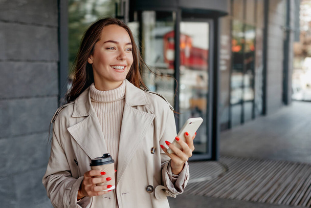 mladá ukrajinská žena používá svůj smartphone, drží šálek kávy a usmívá se na pozadí kancelářské budovy. Studentka odpočívá poblíž budovy univerzity a pije kávu - Fotografie, Obrázek