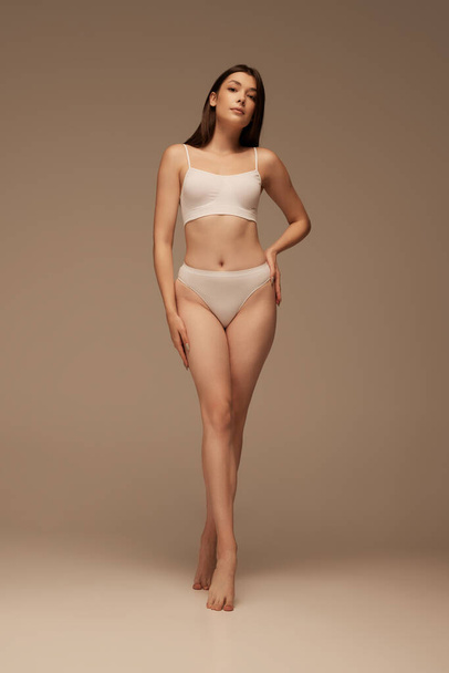 Full-length portrait of young slim woman posing in white underwear isolated over dark beige studio background. Concept of beauty, body and skin care, health, plastic surgery, cosmetics, sport - Fotó, kép