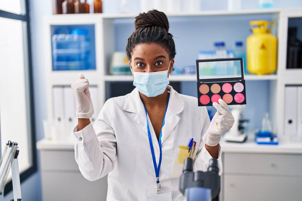 Jeune afro-américain avec des tresses travaillant au laboratoire scientifique faire maquillage crier fier, célébrer la victoire et le succès très excité avec les bras levés  - Photo, image