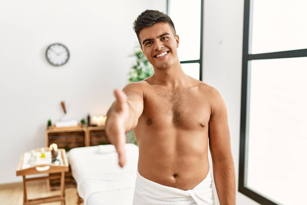 Joven hombre hispano de pie sin camisa en el centro de spa sonriendo amable ofreciendo apretón de manos como saludo y bienvenida. negocios exitosos.  - Foto, Imagen