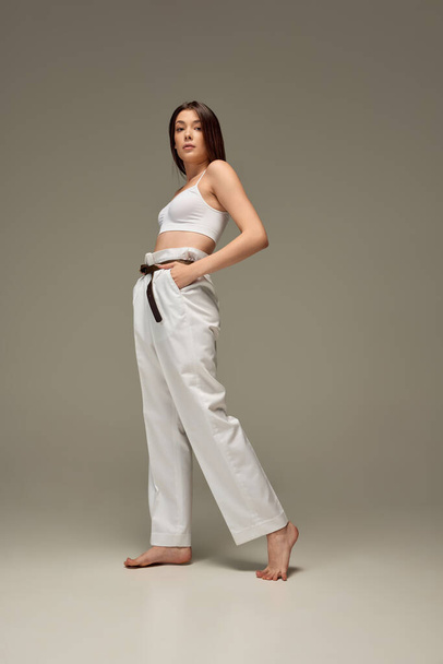Full-length portrait of young beautiful woman posing in white casual outfit of top and pants isolated over grey studio background. Concept of beauty, fashion, style, body and skin care, cosmetics - Fotoğraf, Görsel