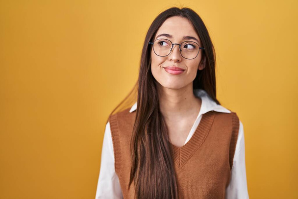 Jeune femme brune debout sur fond jaune portant des lunettes souriant regardant vers le côté et regardant loin en pensant.  - Photo, image