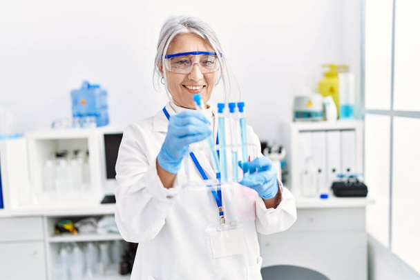 Mujer de pelo gris de mediana edad que usa uniforme científico usando un portátil que sostiene el tubo de ensayo en el laboratorio - Foto, Imagen