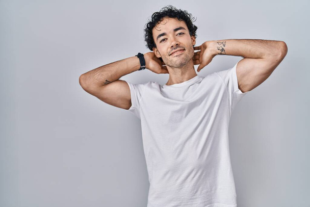 Spaanse man staat over geïsoleerde achtergrond ontspannen en strekken, armen en handen achter hoofd en nek glimlachend gelukkig  - Foto, afbeelding