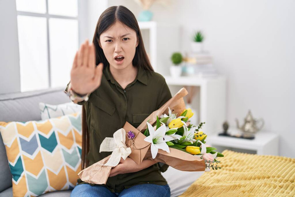 Китайська молода жінка, що тримає букет білих квітів робить зупинки жест пальцями, гнів і розчарування вираження.  - Фото, зображення
