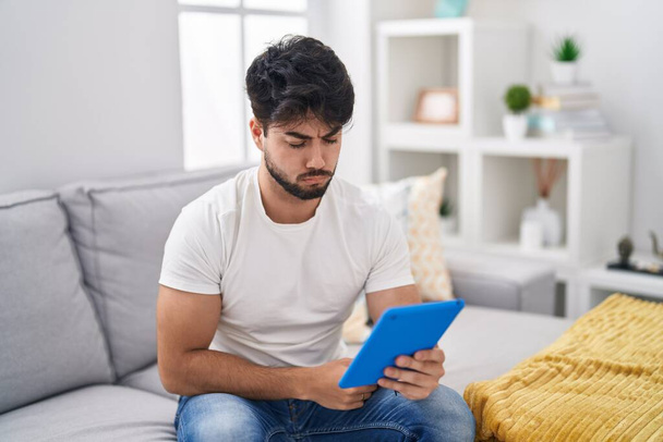 Hispanic man with beard using touchpad sitting on the sofa skeptic and nervous, frowning upset because of problem. negative person.  - Photo, Image