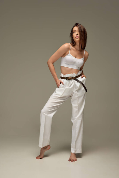 Full-length portrait of young beautiful woman posing in white casual outfit of top and pants isolated over grey background. Comfort. Concept of beauty, fashion, style, body and skin care, cosmetics - Fotó, kép