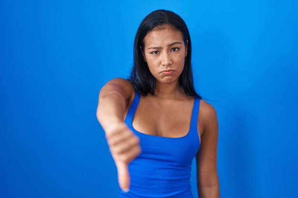 Mujer hispana de pie sobre fondo azul mirando infeliz y enojado mostrando rechazo y negativo con los pulgares hacia abajo gesto. mala expresión.  - Foto, Imagen
