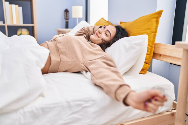 Молода іспаномовна жінка розтягує руки лежачи на ліжку в спальні
 - Фото, зображення