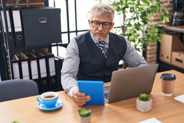 Middelbare leeftijd grijs-harige man zakenman met behulp van laptop en touchpad op kantoor - Foto, afbeelding