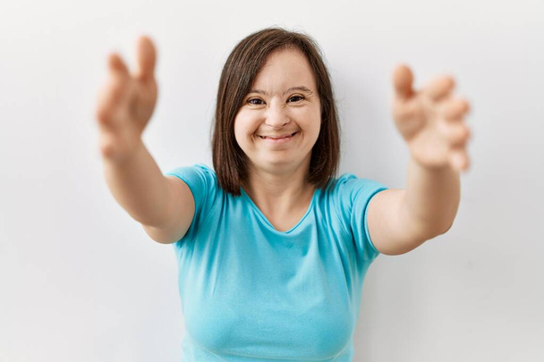 Junge Frau mit Down-Syndrom, die vor isoliertem Hintergrund steht und mit offenen Armen in die Kamera lächelt, um sich zu umarmen. Fröhlicher Ausdruck des Glücks.  - Foto, Bild