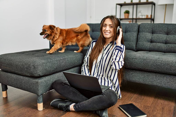 Young hispanic woman using laptop sitting on floor with dog at home - Photo, Image
