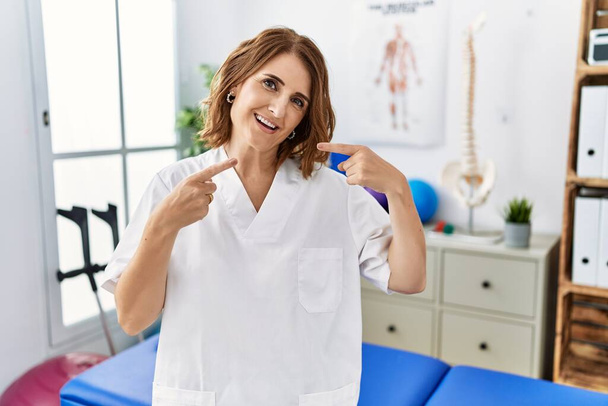 Középkorú fizioterapeuta nő dolgozik fájdalom helyreállítási klinika mosolyogva vidám bemutató és rámutatva ujjal fogak és száj. fogászati egészség fogalma.  - Fotó, kép