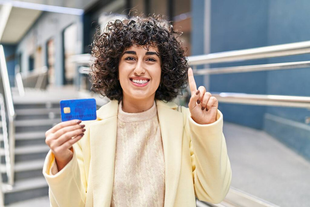 Nuori brunette nainen kihara tukka tilalla luottokortti hymyillen idea tai kysymys sormella onnellinen kasvot, numero yksi  - Valokuva, kuva