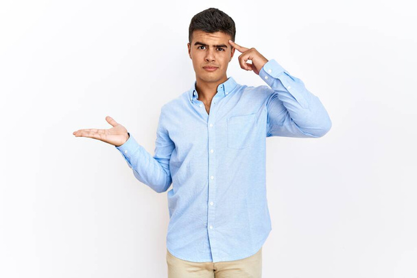 孤立した背景の上に立ったビジネスシャツを着た若いヒスパニック系の男が混乱し、コピースペースを示し、額に指を指すオープン手のひらに悩まされました。考えてみてください.  - 写真・画像