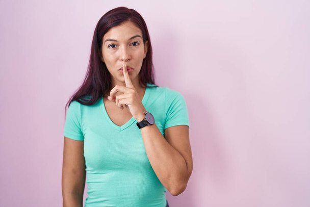 ピンクの背景の上に立つ若いヒスパニック系の女性が唇に指で静かにするように求めている。沈黙と秘密の概念.  - 写真・画像