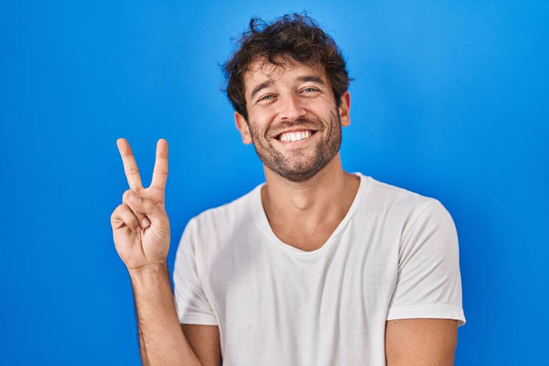 Ισπανόφωνος νεαρός άνδρας που στέκεται πάνω από το μπλε φόντο χαμογελώντας με χαρούμενο πρόσωπο να κλείνει το μάτι στην κάμερα κάνοντας σήμα νίκης με τα δάχτυλα. Νούμερο δύο..  - Φωτογραφία, εικόνα