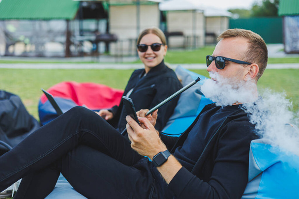 Молодая женщина и мужчина сидят в парке на бобовом мешке и курят кальян. Счастливая пара отдыхает на открытом воздухе с кальяном - Фото, изображение