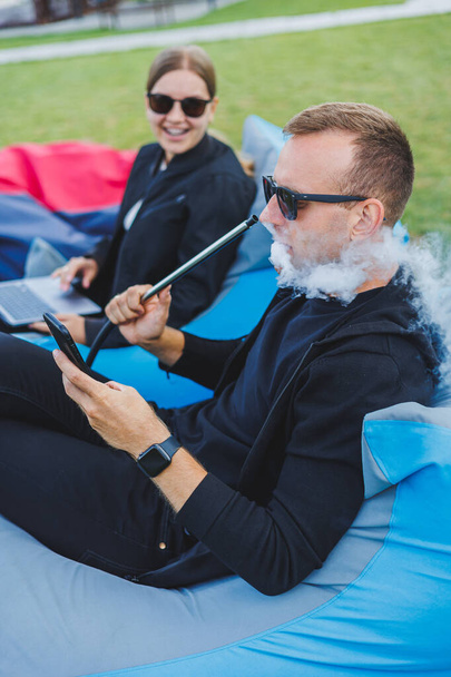 Una joven y un hombre están sentados en un parque en una bolsa de frijoles y fumando narguile. Pareja feliz relajándose al aire libre con narguile - Foto, imagen