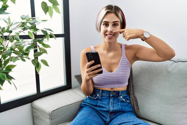 Νεαρή όμορφη γυναίκα που χρησιμοποιεί το μήνυμα πληκτρολόγησης smartphone κάθεται στον καναπέ δείχνοντας με το χέρι το δάχτυλο στο πρόσωπο και τη μύτη, χαμογελώντας χαρούμενα. έννοια ομορφιά  - Φωτογραφία, εικόνα