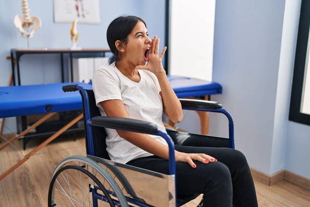 Giovane donna ispanica seduta sulla sedia a rotelle alla clinica di fisioterapia incapace e confusa con le braccia aperte, nessuna idea e viso dubbioso.  - Foto, immagini