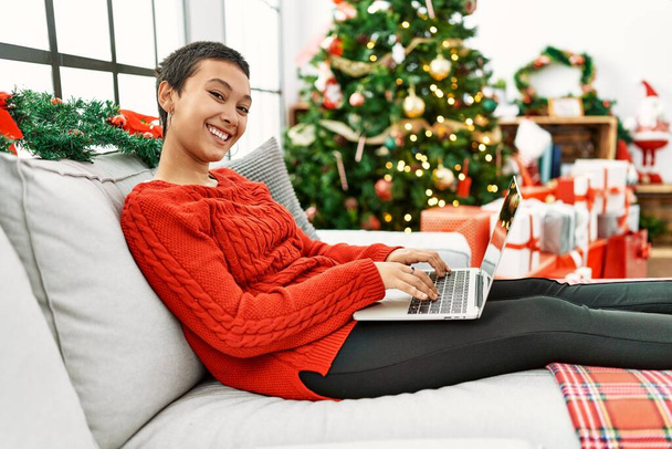 Young hispanic woman using laptop sitting by christmas tree at home - Zdjęcie, obraz
