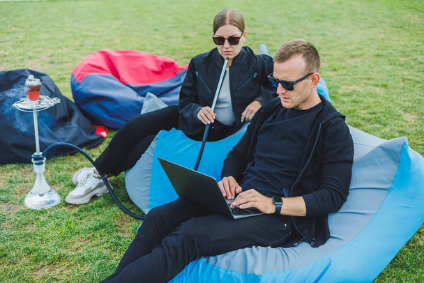 Ein junges Paar entspannt sich im Park und raucht Wasserpfeife. Ein Mann und eine Frau sitzen in der Natur auf Stühlen und rauchen eine Wasserpfeife. Familienurlaub - Foto, Bild