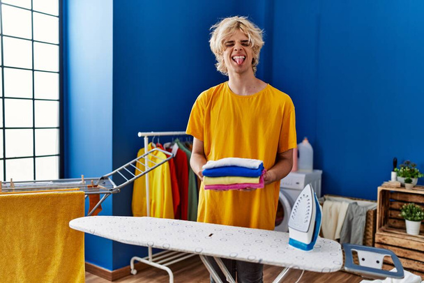 Young man ironing clothes at home sticking tongue out happy with funny expression.  - Foto, Imagen
