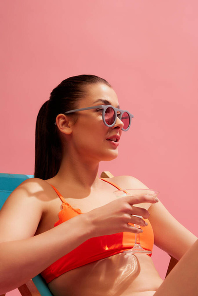 Portrait of young beautiful woman posing in swimming suit and sunglasses isolated over pink background. Concept of beauty, summer season, cosmetology, swimwear collection, body and skin care - Photo, Image