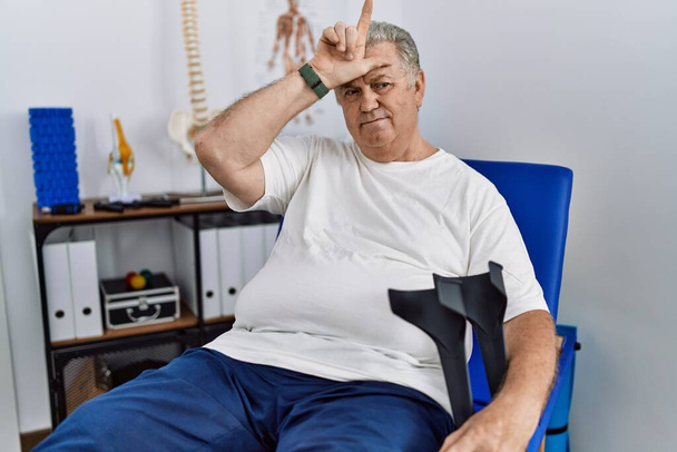 Uomo anziano caucasico alla clinica di fisioterapia che tiene le stampelle prendendo in giro le persone con le dita sulla fronte che fanno gesti da perdente deridendo e insultando.  - Foto, immagini