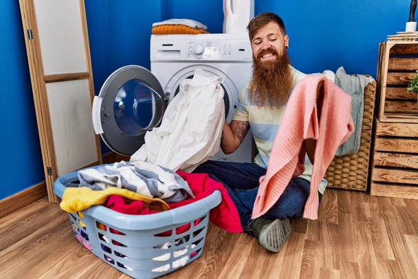 Joven pelirroja sonriendo confiado lavando ropa en la lavandería - Foto, Imagen