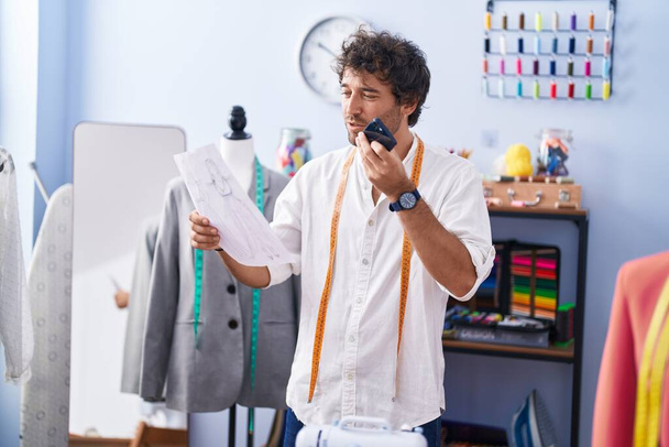 Νεαρός Ισπανός ράφτης μιλάει σε smartphone που αναζητούν σχεδιασμό ρούχων στο εργοστάσιο ρούχων - Φωτογραφία, εικόνα