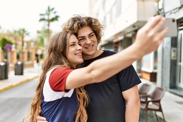 Joven pareja caucásica sonriendo feliz y abrazando hacer selfie por el teléfono inteligente en la ciudad. - Foto, Imagen