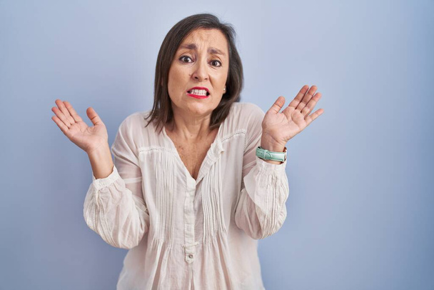 Middle age hispanic woman standing over blue background clueless and confused expression with arms and hands raised. doubt concept.  - Photo, Image