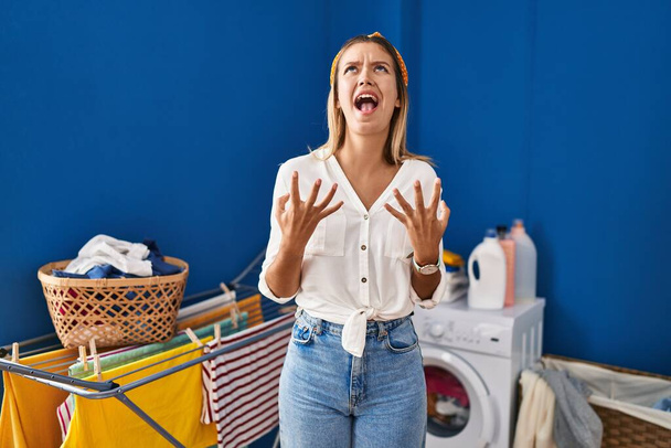 Young blonde woman at laundry room crazy and mad shouting and yelling with aggressive expression and arms raised. frustration concept.  - Foto, Imagem