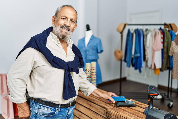 Старший седой мужчина платит кредитной картой и телефоном в магазине одежды - Фото, изображение