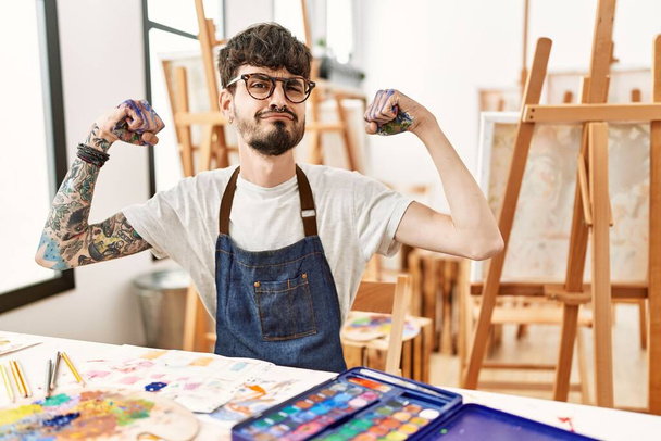 Homme hispanique avec barbe au studio d'art montrant les muscles des bras souriant fier. concept de fitness.  - Photo, image