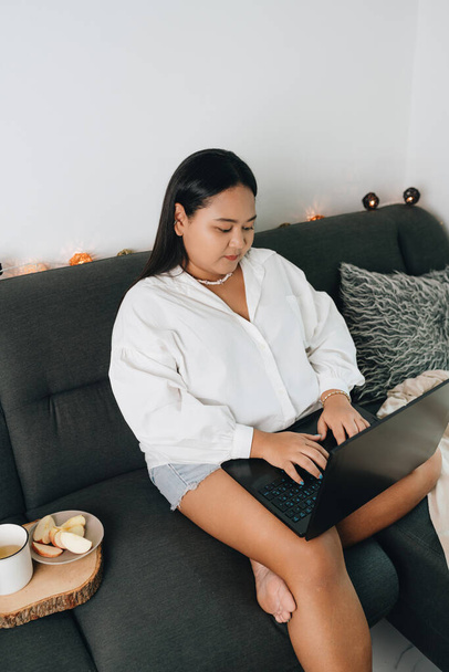 Portrait of a young female freelancer sitting on the couch and using laptop at home - 写真・画像