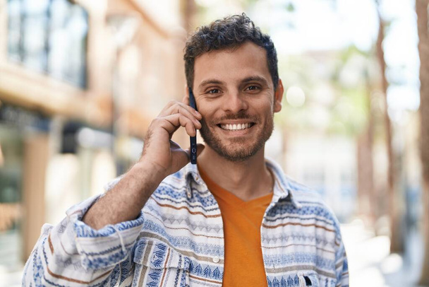 Junger hispanischer Mann lächelt selbstbewusst und spricht auf der Straße mit dem Smartphone - Foto, Bild